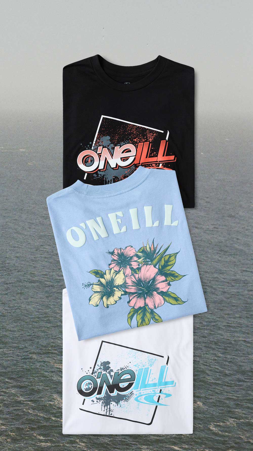 Three folded Oneill t-shirts displayed against a blurred water background: a black shirt with a red and white graphic, a light blue shirt with pink and yellow flowers and white 'ONEILL' text, and a white shirt with a blue and black graphic.