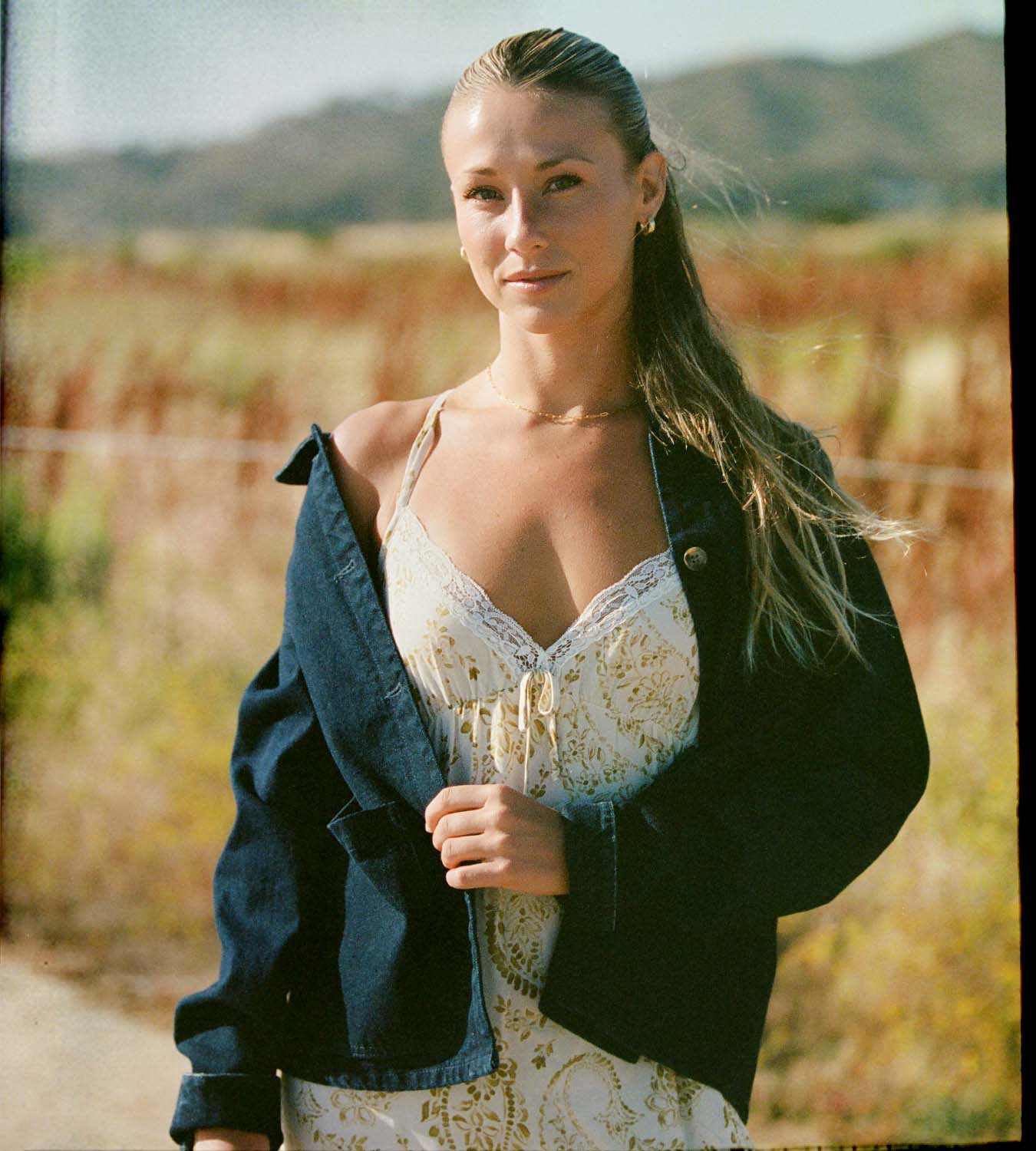 A woman standing outdoors in a field, wearing a white lace-trimmed camisole dress with a cream floral pattern, a dark blue denim jacket draped off her shoulders, and a gold necklace.