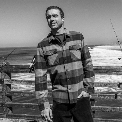 Man wearing a plaid shirt standing by a wooden fence with ocean in the background