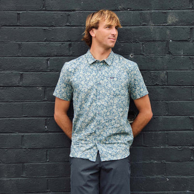 A man with blond hair wearing a short-sleeve button-up shirt with a blue and white floral pattern, standing against a dark gray brick wall.