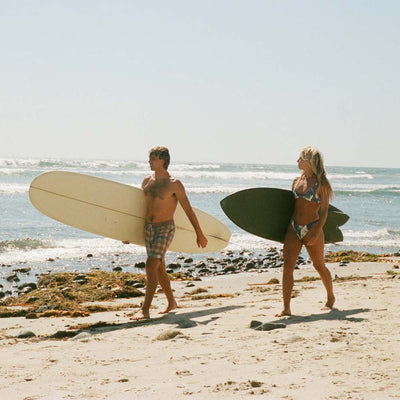 A man and a woman walking on a sandy beach carrying surfboards, with the ocean and pale sky in the background.