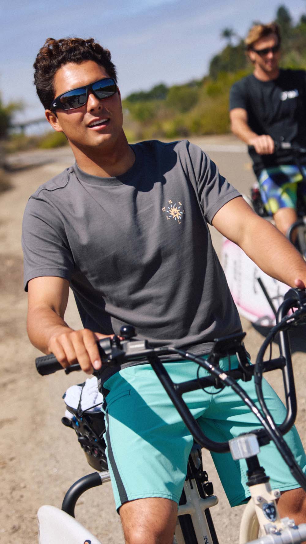 A man riding a bicycle outdoors wearing a dark gray crewneck t-shirt with a small sun graphic on the left chest, paired with teal shorts. Another cyclist is visible in the background on a sunny day.