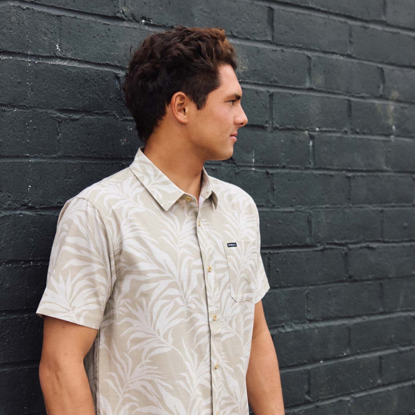 A man wearing a short-sleeve button-up shirt with a light beige and white palm leaf pattern, standing against a dark gray brick wall.