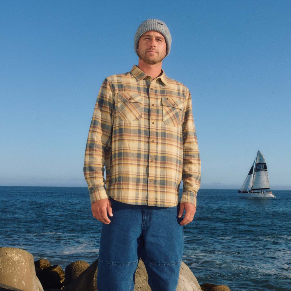A man wearing a beige and brown plaid flannel shirt, blue jeans, and a gray beanie stands on a rocky shoreline with the ocean and a sailboat in the background.