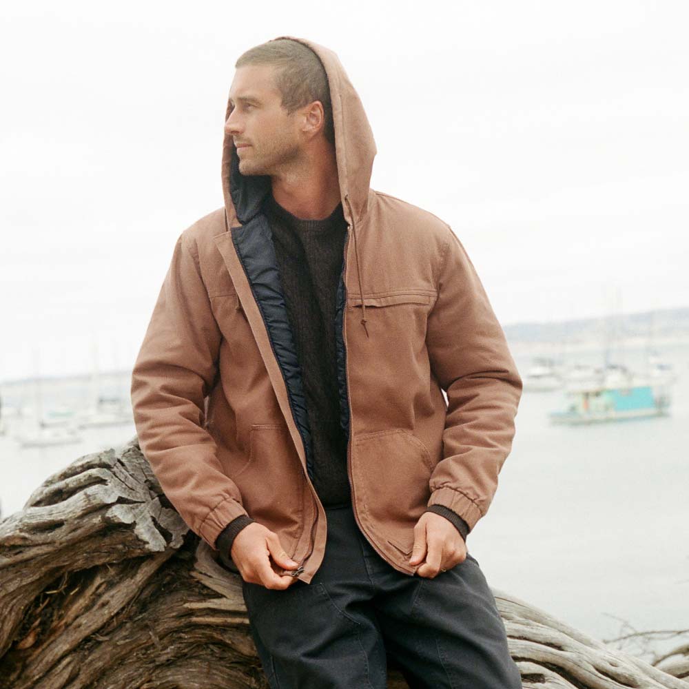A man wearing a brown hooded jacket over a black shirt and dark pants, sitting on a large piece of driftwood with a harbor and boats in the background.