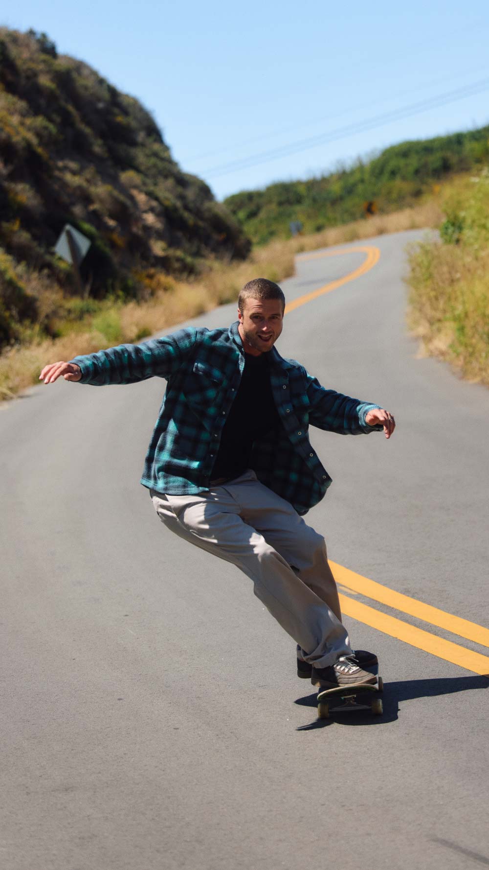 A man riding a skateboard down a paved road with yellow double lines, wearing a green and black plaid flannel shirt, a black t-shirt, and light gray sweatpants.