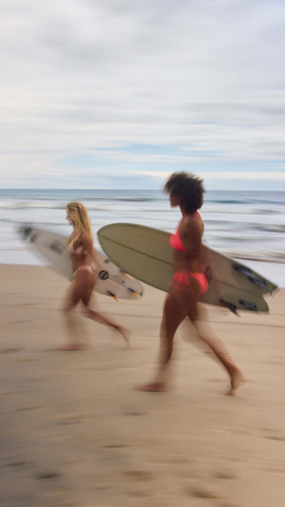 Two women carrying surfboards run along a sandy beach under an overcast sky. One woman wears a bright pink bikini and the other wears a beige bikini.