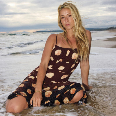 A woman kneeling in shallow ocean water on a beach, wearing a brown spaghetti-strap dress with a beige seashell pattern.