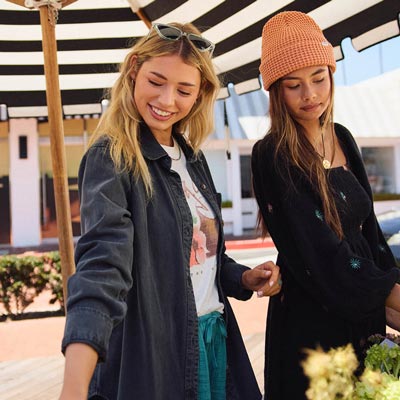 Two women standing outdoors under a striped canopy, one wearing a denim jacket and sunglasses on her head, the other in a black outfit with a pink beanie.