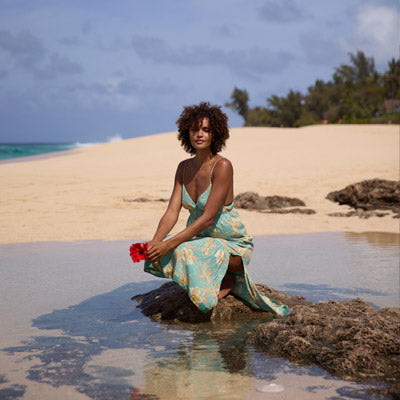Woman in a floral dress sitting on a beach holding a red flower.