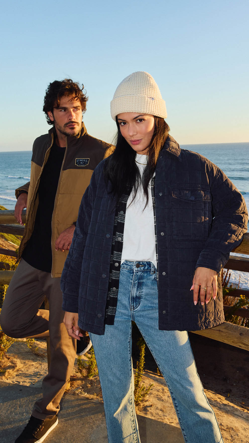 Two people standing on a beach with ocean view, wearing jackets and hats.