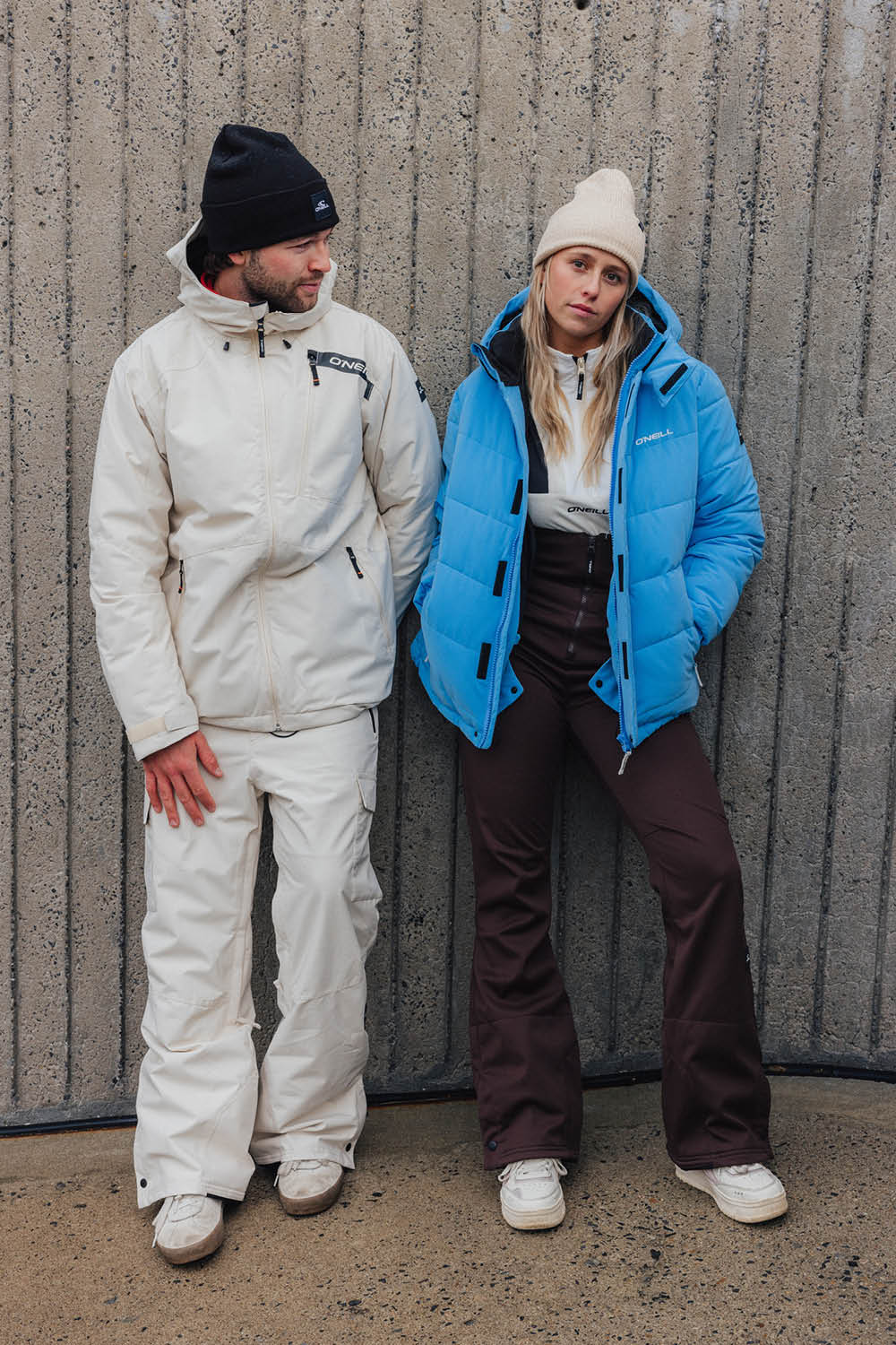 Two people wearing winter jackets and pants standing against a textured wall.