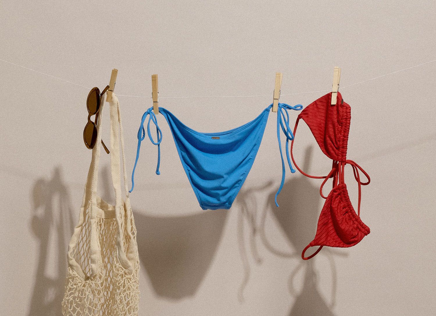 Blue and red bikini bottoms hanging on a line with a beige bag and sunglasses.