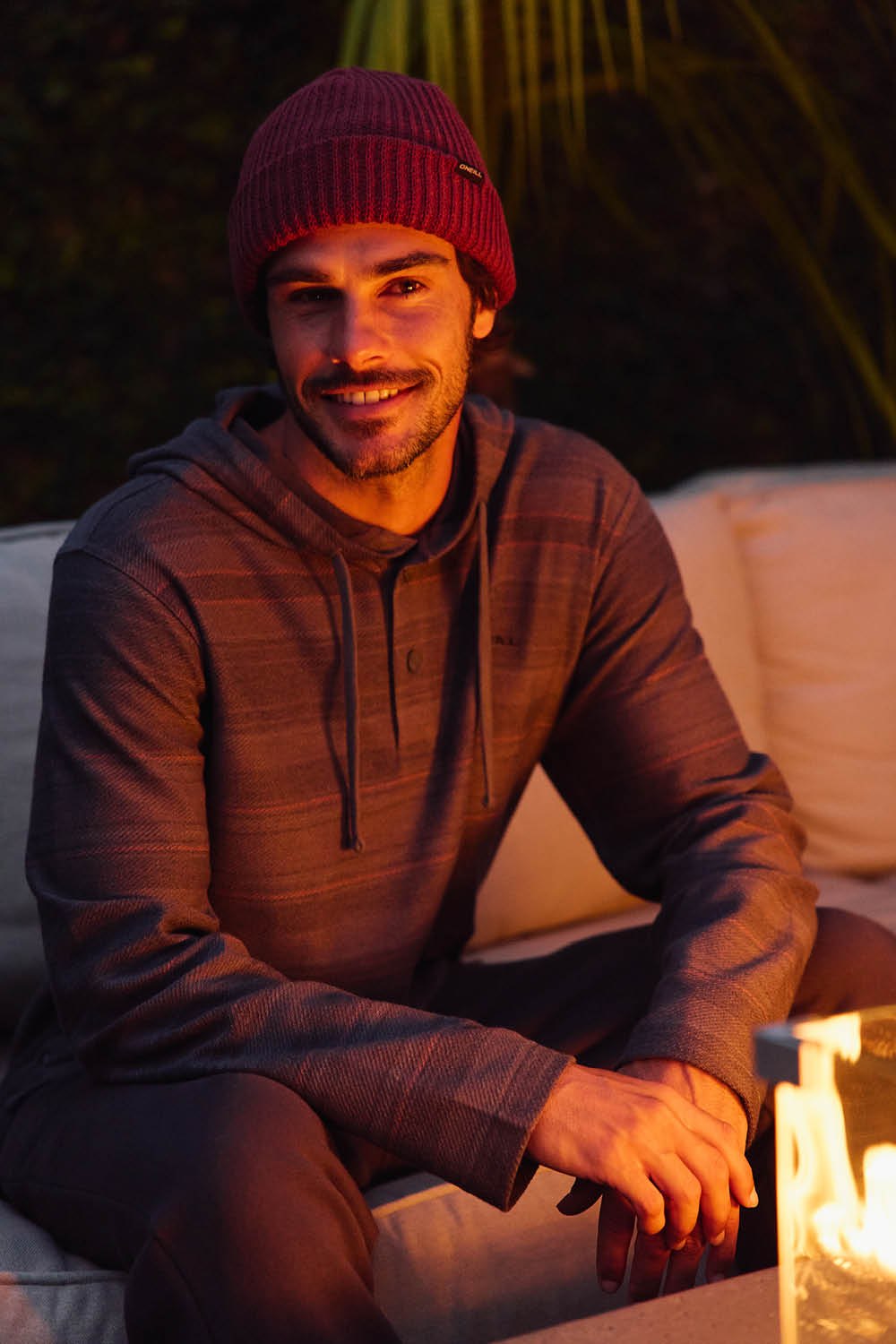 Man wearing a red beanie and brown hoodie sitting by a fire outdoors.