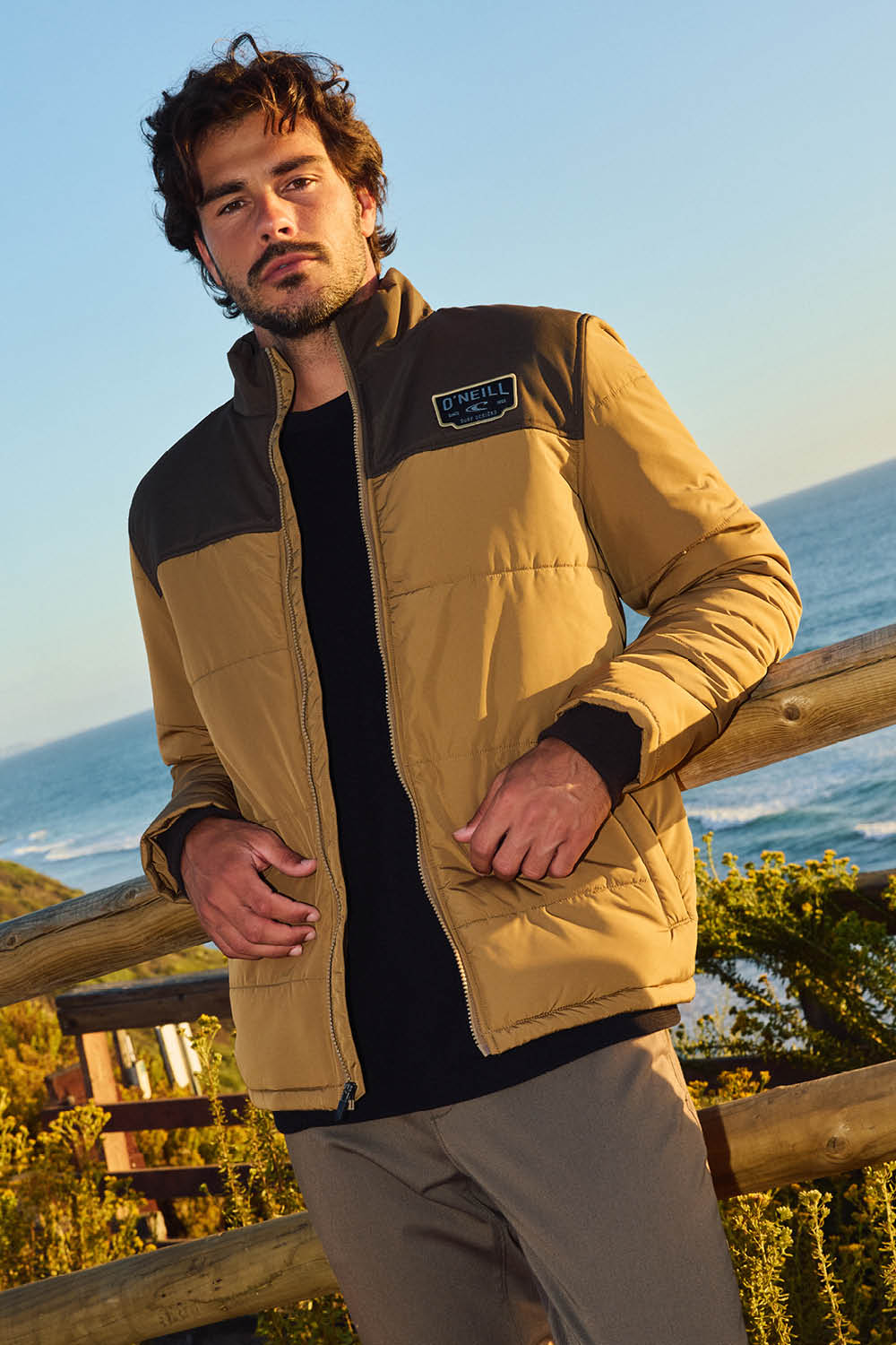 Man wearing a beige and black jacket standing outdoors with ocean view