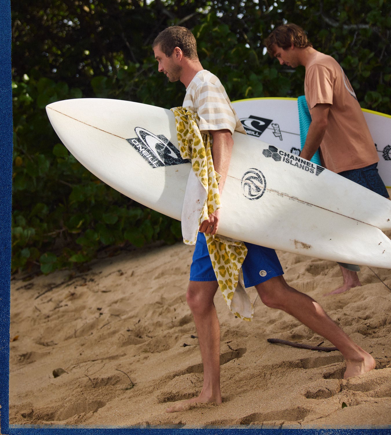 Two men walking on a sandy beach carrying surfboards with a tropical background.