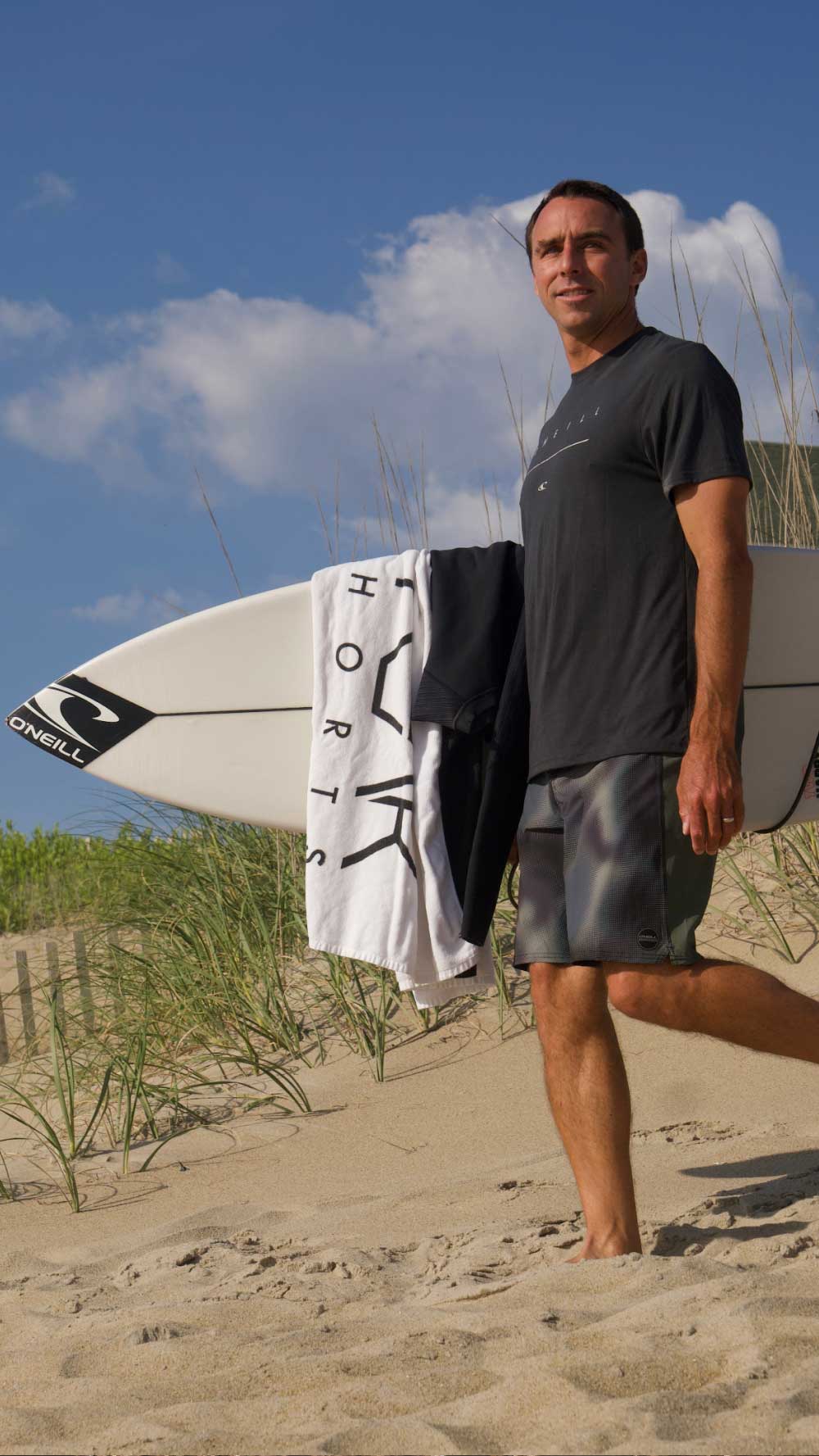 A man walking on a sandy beach carrying a white surfboard under his arm, with a white and black surf towel draped over the board. He is wearing a dark gray t-shirt and patterned swim trunks, with dunes, grass, and a blue sky in the background.