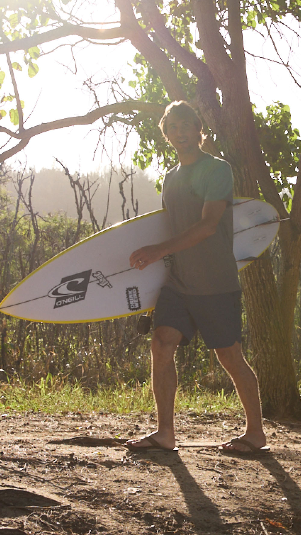 Person holding a surfboard in a natural setting with trees and sunlight.