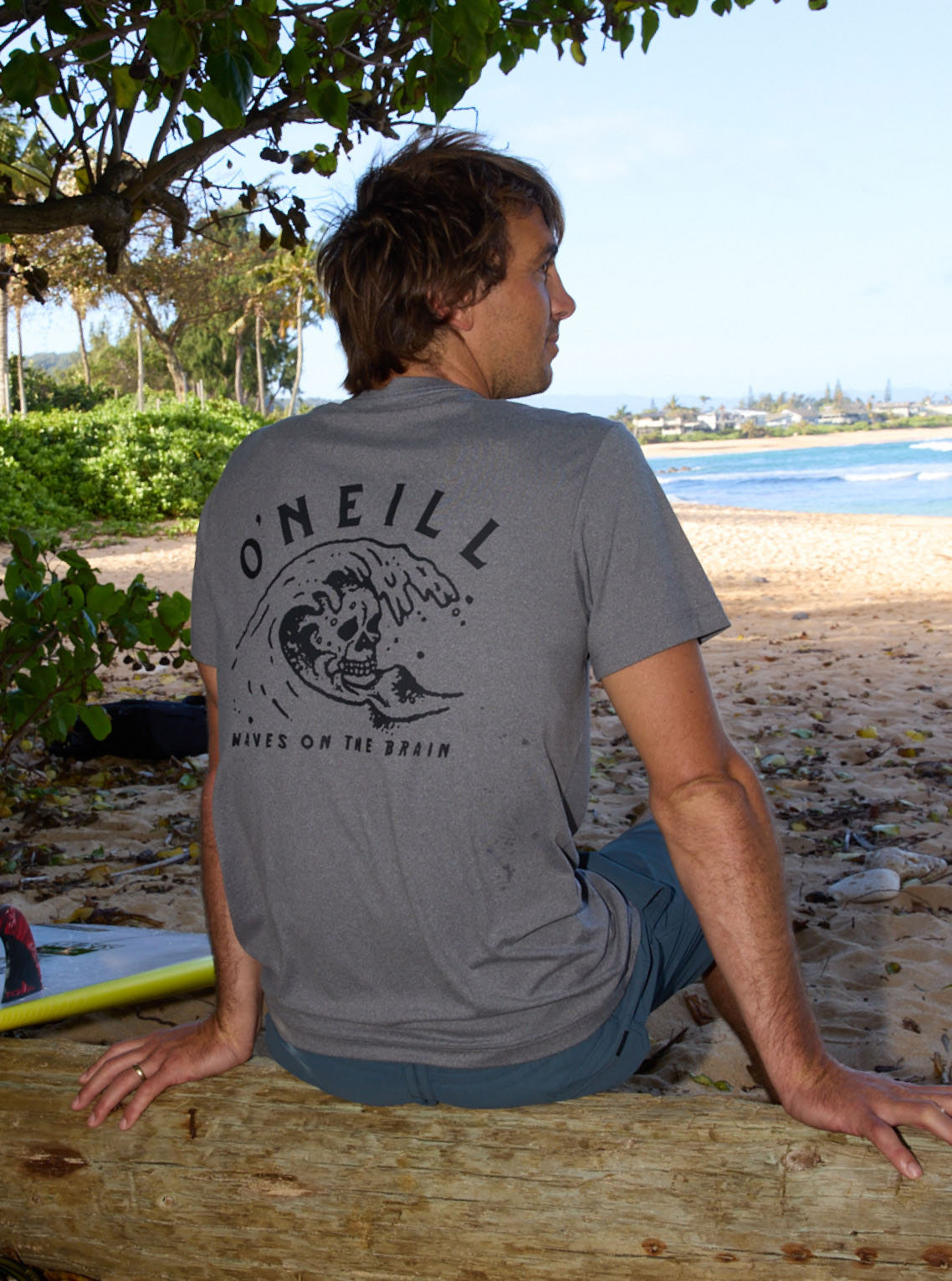 Man wearing a gray O'Neill t-shirt sitting on a log by a beach.