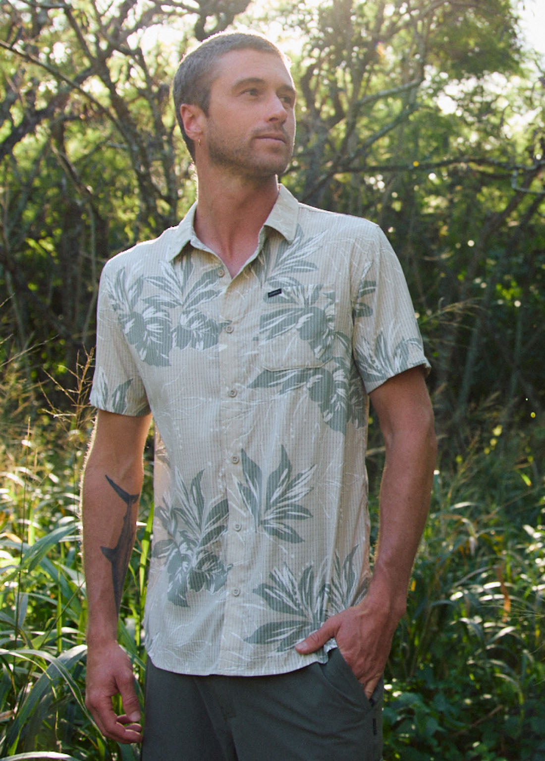 Man wearing a patterned shirt standing in a natural setting with trees and greenery.
